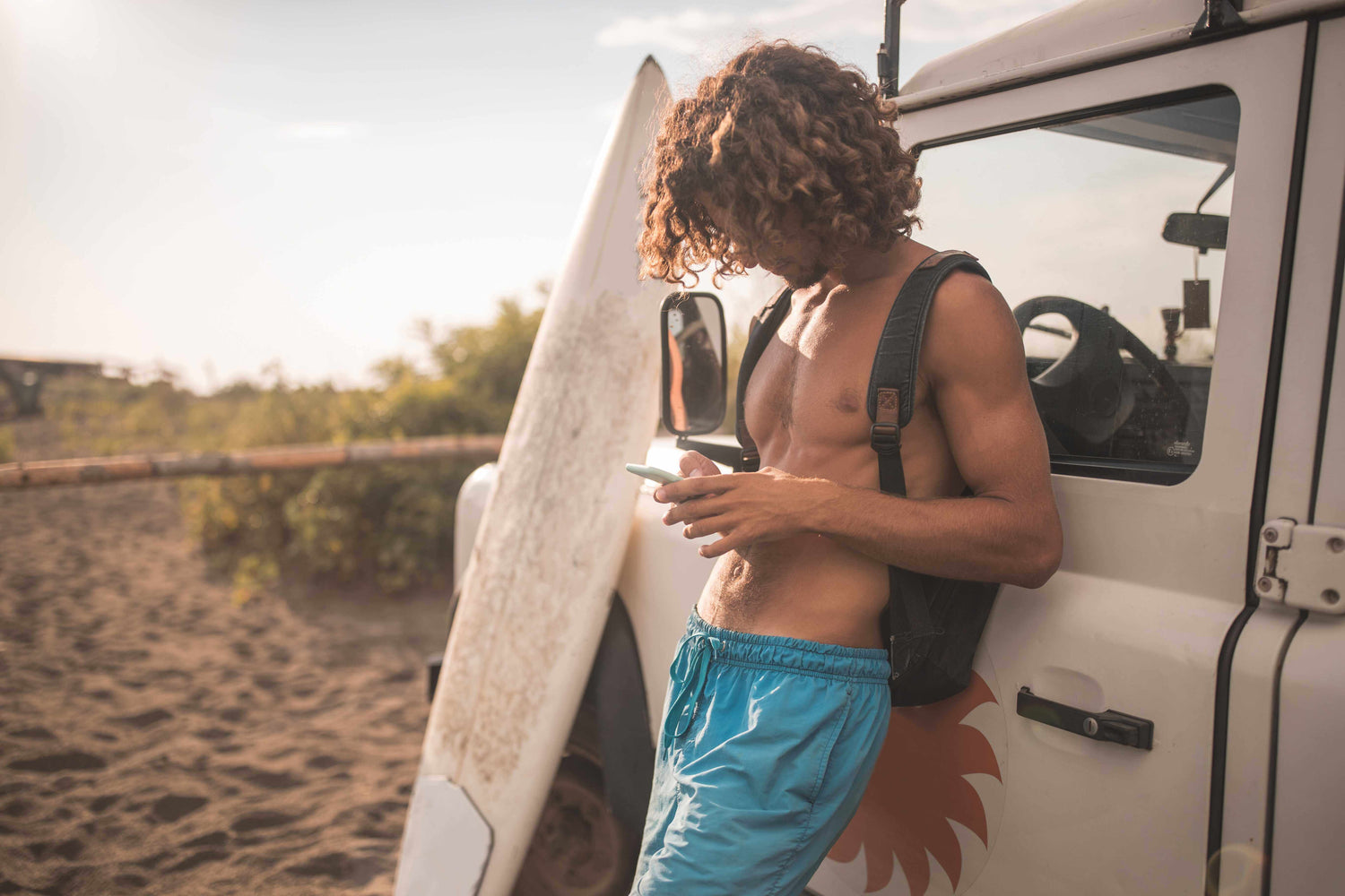 Man with a surfboard leaning against a vehicle on a beach