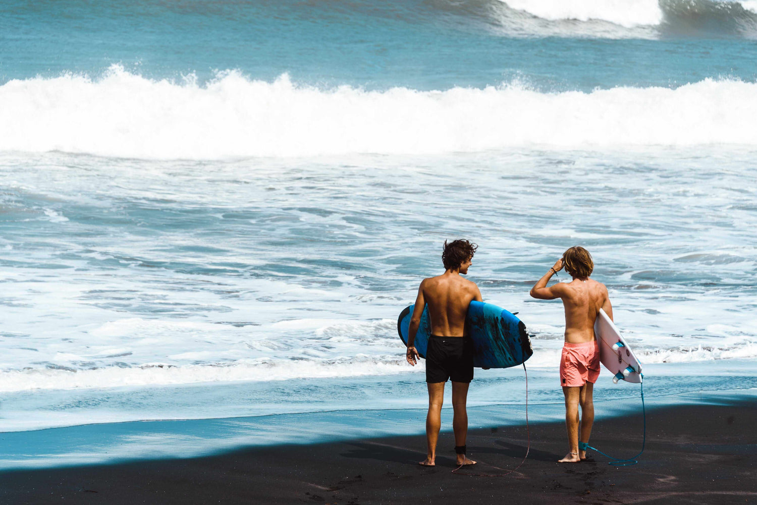 Two surfers with their boards standing on a beach facing the ocean.