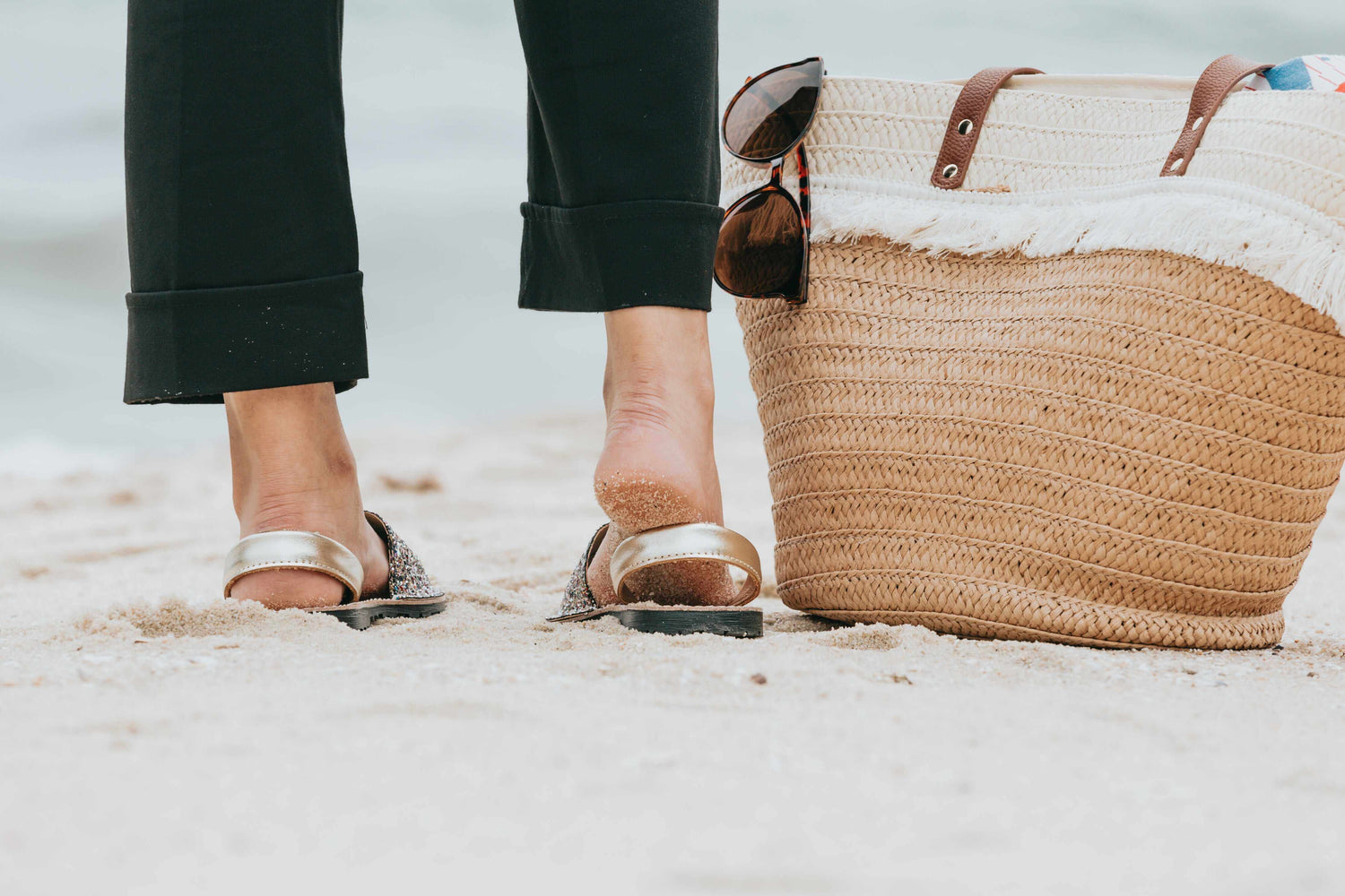 Person wearing sandals on a sandy surface with a straw bag and sunglasses.