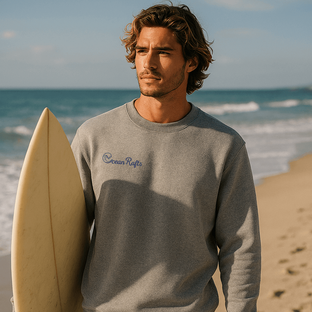 Man holding a surfboard on a beach wearing a gray sweatshirt with 'Ocean Rats' logo.